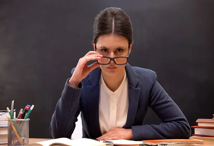 prof en colère assise à son bureau en classe et fixant droit devant elle en abaissant ses lunettes